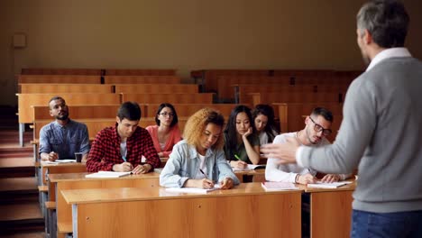 Professor-liest-Vortrag-zur-Gruppe-der-gemischtrassigen-Schüler-im-Klassenzimmer-mit-Papieren-stehen-und-sprechen,-während-junge-Menschen-ihn-hören-und-schreiben.