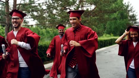 Slow-motion-of-cheerful-grads-running-together-under-rain-waving-diplomas-and-laughing-wearing-red-gowns-and-mortar-boards.-Small-rain-is-visible.