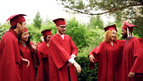 Begeisterte-junge-Männer-und-Frauen-Absolventen-feiern-Abschluss-Tag-lachen,-dabei-hohe-fünf,-umarmt-und-tanzen-außerhalb-auf-dem-Campus.-Menschen-tragen-traditionelle-Kleidung.