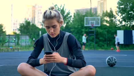 Beautiful-young-woman-sitting-on-basketball-court-and-typing-on-phone,-wearing-earphones,-park-with-skyscrapers-in-background