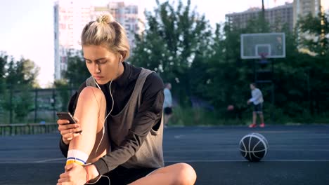 Charmante-glücklich-weiblichen-Basketball-Spieler-tragen-Kopfhörer-und-tippen-auf-Telefon,-sitzen-auf-Boden,-Männer-spielen-im-Hintergrund