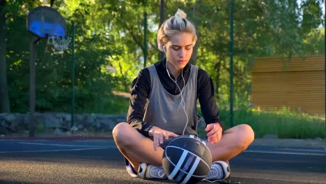 Charming-young-female-basketball-player-standing-up-from-ground-and-going-away,-holding-ball,-basketball-court-in-open-air,-sunset