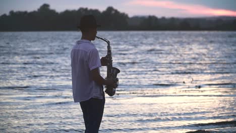 Chico-joven-guapo-es-jugar-el-saxophone-en-la-orilla-del-río