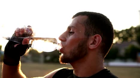 Side-view-of-a-young-male-boxer-in-black-bandages-has-a-rest-during-the-training-outdoors,-drinking-water-from-a-plastic-bottle.-Sun-shines-on-the-background