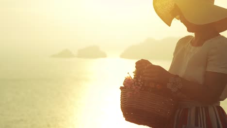 Beautiful-young-woman-wearing-fashion-colorful-dress-with-skirt-and-hat-picking-with-flowers-in-basket-at-sunset-on-Ponza-Island-mountain-coast-Italy.