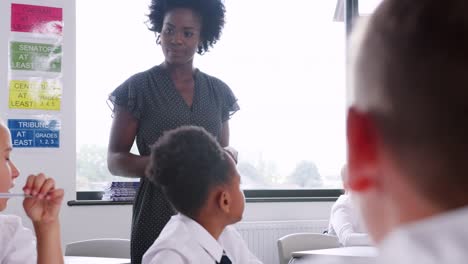 Female-High-School-Tutor-Teaching-Group-Of-Students-Wearing-Uniform-Working-Around-Table-In-Classroom