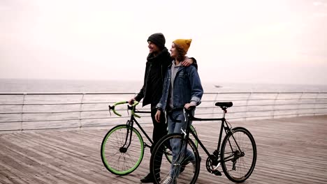 Hermosa-pareja-sonriente-de-los-hipsters-jóvenes-caminando-juntos-abrazando-con-sus-bicicletas-cerca-del-mar-en-día-de-otoño.-Chica-joven-de-sombrero-amarillo-en-algo.-Caminando-por-cubierta-de-madera-durante-el-día.-Horizonte-de-la-mar