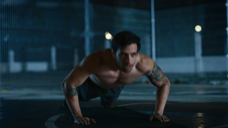 Strong-Muscular-Fit-Shirtless-Young-Man-is-Doing-Push-Up-Exercises.-He-is-Doing-a-Workout-in-a-Fenced-Outdoor-Basketball-Court.-Night-Footage-After-Rain-in-a-Residential-Neighborhood-Area.