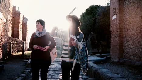 Young-beautiful-female-excursion-tour-guide-showing-historic-antique-streets-of-Pompeii,-Italy-to-a-senior-woman-tourist