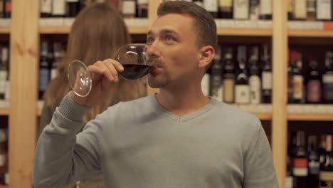 Handsome-pensive-guy-with-a-wine-glass-in-a-liquor-store.-Woman-in-the-background-is-choosing-a-wine-bottle.