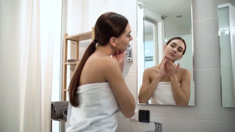 Cuidado-de-la-piel.-Mujer-tocando-la-cara-y-mirando-en-el-espejo-en-el-baño