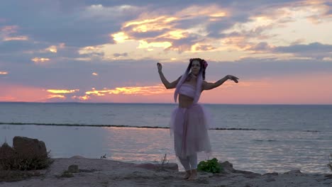 Linda-chica-con-maquillaje-brillante-en-un-vestido-rosa-bailando-en-la-orilla-del-río.-El-baile-de-una-mujer-sensual-con-un-peinado-maravilloso-con-flores.-Cámara-lenta.