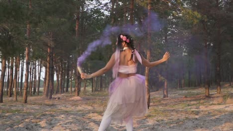 Charming-woman-with-bright-makeup-in-a-pink-dress-dancing-with-smoke-bombs-on-the-background-of-pine-trees.-The-dance-of-a-sensual-girl-with-a-wonderful-hairstyle-with-flowers.-Slow-motion.