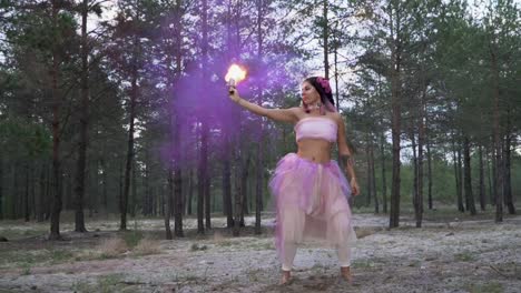 Cute-young-woman-with-bright-makeup-in-a-pink-dress-dancing-with-burning-smoke-bombs-on-the-background-of-pine-trees.-The-dance-of-a-sensual-girl-with-a-flower-hairstyle.-Slow-motion.