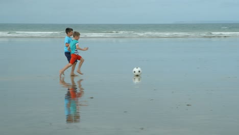 Two-boys-laughing-and-sprinting-along-a-beach-to-get-to-their-soccer-ball.