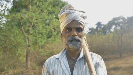 A-male-farmer-standing-in-his-agricultural-land-surrounded-by-trees-carrying-pickax.