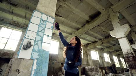 Attractive-girl-urban-artist-is-painting-graffiti-in-abandoned-building-with-dirty-walls-and-windows,-she-is-using-paint-spray.-Modern-artwork-and-creative-people-concept.