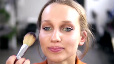 Vista-frontal-de-una-hermosa-modelo-mujer-sonriente-durante-el-proceso-de-maquillaje.-Maquillaje-artista-termina-el-trabajo,-poniendo-un-polvo-con-un-cepillo.-Imágenes-de-cerca