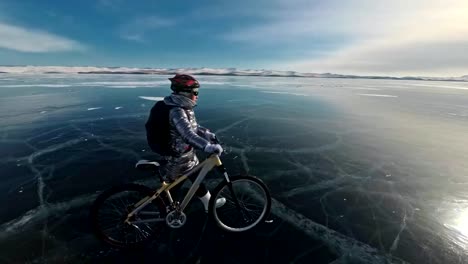 Mujer-está-caminando-al-lado-de-la-bicicleta-en-el-hielo.-La-chica-está-vestida-con-un-plateado-abajo-chaqueta,-mochila-y-casco.-Hielo-del-lago-Baikal-congelado.-Los-neumáticos-de-la-bicicleta-están-cubiertos-con-los-puntos-especiales.