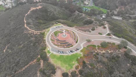 Amazing-Cinematic-Drone-Shot-Dropping,-Flying-Backwards,-and-Panning-Up-Over-Mt.-Soledad-in-San-Diego,-California-to-Reveal-the-Hills-and-Water-in-the-Distance.