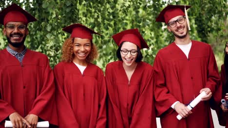 Dolly-shot-slow-motion-of-laughing-young-people-graduates-holding-diplomas-standing-in-line-outdoors-on-campus-enjoying-celebration.-Graduation-day-and-youth-concept.