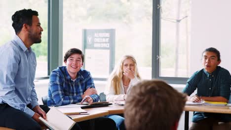 Male-High-School-Tutor-Sitting-On-Desk-Teaching-Class