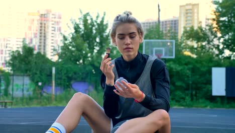 Attractive-female-basketball-player-sitting-on-court-and-doing-make-up,-park-and-buildings-in-background