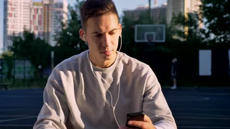 Confident-young-man-typing-on-phone,-listening-music-through-earphones-and-looking-at-camera,-people-playing-in-background