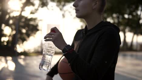Down-up-footage-of-a-young-sporty-girl-basketball-player-have-a-rest-after-training-on-the-local-outdoors-court,-drinking-a-water-from-a-plastic-bottle.-Blurred-view-of-a-morning-sunlight-and-trees-on-the-background