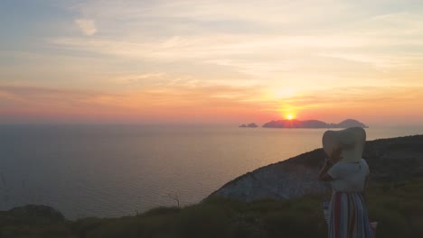 Hermosa-mujer-joven-con-sombrero-de-moda-colorido-vestido-con-la-falda-y-flores-mirando-la-puesta-de-sol-en-montaña-de-la-isla-de-Ponza-Italia.-Tiro-de-Drone-aéreo.