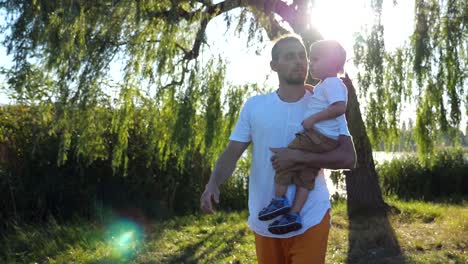 Junger-Vater-seinen-kleinen-Sohn-in-den-Händen-halten-und-im-Sommerpark-an-einem-sonnigen-Tag-zu-Fuß.-Glückliche-Familie-Zeit-miteinander-zu-verbringen-und-die-Natur-genießen.-Schöne-Landschaft-im-Hintergrund.-Slow-Motion-hautnah