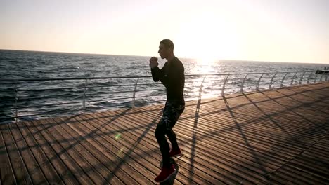 Male-boxer-fighter-training-punches-outdoors,-workout-with-invisible-opponent.-Healthy-lifestyle-and-energy-with-motivation---concept-of-strength-training-near-the-sea.-Morning-sun-shines-on-the-background