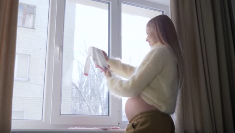 Waiting-of-baby,-young-pregnant-female-with-big-tummy-looks-at-little-children-clothes-for-future-baby-against-window-in-sunlight