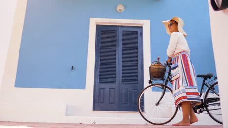 Mujer-joven-feliz-con-bicicleta-caminando-frente-a-patio-de-la-puerta-de-la-casa-azul.-Camisa-de-moda-blanco,-gran-sombrero,-falda-colorida-y-gafas-de-sol.-Isla-de-Ponza,-Italia.