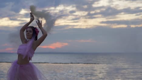 Linda-chica-con-maquillaje-brillante-en-un-vestido-rosado-bailando-con-bombas-de-humo-en-la-orilla-del-río.-El-baile-de-una-mujer-sensual-con-un-peinado-maravilloso-con-flores.-Cámara-lenta.