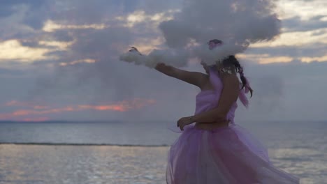 Tierna-chica-con-maquillaje-brillante-en-un-vestido-rosado-bailando-con-bombas-de-humo-en-la-orilla-del-río.-El-baile-de-una-mujer-sensual-con-un-peinado-maravilloso-con-flores.-Cámara-lenta.