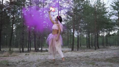 Cute-tender-woman-with-bright-makeup-in-a-pink-dress-dancing-with-burning-smoke-bombs-on-the-background-of-pine-trees.-The-dance-of-a-sensual-girl-with-a-flower-hairstyle.-Slow-motion.