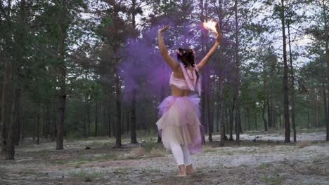 Mujer-tierna-linda-con-maquillaje-brillante-en-un-vestido-rosado-bailando-con-quema-de-bombas-de-humo-en-el-fondo-de-los-pinos.-El-baile-de-una-chica-sensual-con-un-peinado-de-flor.-Cámara-lenta.