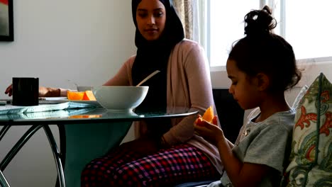 Madre-joven-usando-laptop-mientras-su-hija-está-comiendo-frutas-en-casa-4k