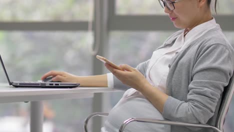 Chinesisch-schwangere-Bürofrau-arbeitet-mit-Laptop-und-Zelle