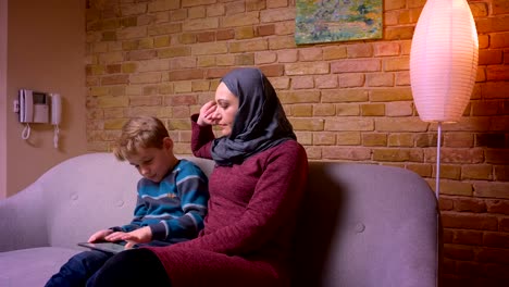 Concentrated-small-boy-playing-game-on-tablet-and-his-muslim-mother-in-hijab-observing-his-activity-at-home.