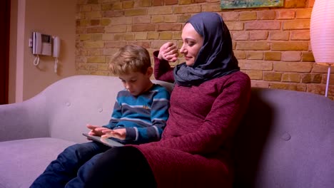 Konzentrierter-kleiner-Junge-spielt-Spiel-auf-Tablet-und-seine-muslimische-Mutter-in-Hijab-streichelt-ihn-zärtlich-zu-Hause.