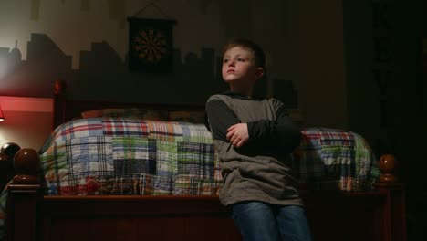 Sad-young-boy-stands-at-the-foot-of-his-bed-with-crossed-arms