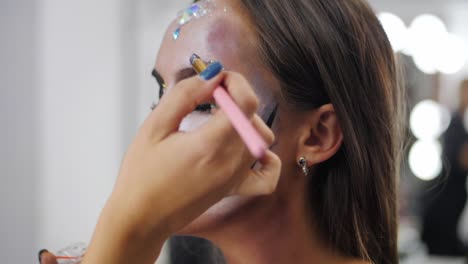 Artista-de-maquillaje-pinturas-de-maquillaje-para-Halloween-en-estudio.-Mujer-dibujando-una-calavera-glamorosa-con-pedrería-y-lentejuelas-en-una-hermosa-joven-con-el-pelo-largo.-Cámara-lenta