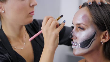 Makeup-artist-paints-greasepaint-for-Halloween-in-studio.-Woman-drawing-a-glamorous-skull-with-rhinestones-and-sequins-on-a-beautiful-young-girl-with-long-hair.-Slow-motion