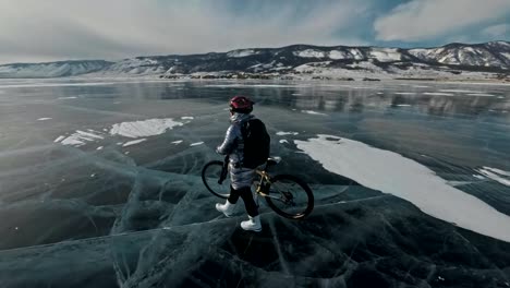 Woman-is-walking-beside-bicycle-on-the-ice.-The-girl-is-dressed-in-a-silvery-down-jacket,-backpack-and-helmet.-Ice-of-the-frozen-Lake-Baikal.-The-tires-on-the-bicycle-are-covered-with-special-spikes.