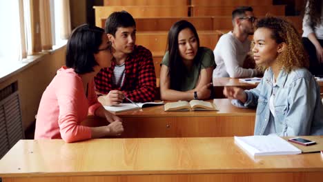 Studierende-diskutieren-Teamprojekt-sitzen-am-Schreibtisch-in-der-Universität,-Mädchen-und-jungen-sprechen,-Notizbücher,-Stifte-und-Smartphones-sind-sichtbar.-Teamwork-und-Kommunikation-Konzept.