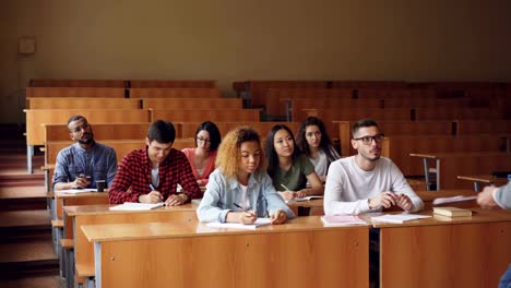 Los-estudiantes-diligentes-son-notas-que-hace-de-la-escritura-en-cuadernos-sentado-en-conferencia-en-la-Universidad-mientras-que-el-profesor-está-hablando-de-pie-delante-de-ellos-en-aulas-grandes.