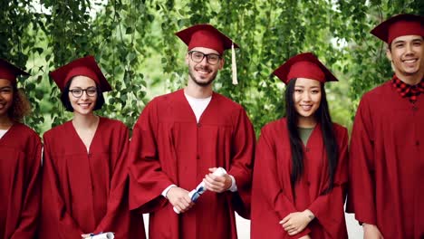 Slow-motion-portrait-of-graduates-in-mortar-boards-and-gowns-looking-at-camera,-smiling-and-holding-diplomas.-Higher-education,-smart-young-people-and-future-concept.