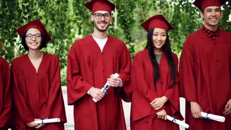 Dolly-shot-of-smiling-graduates-standing-in-line-on-campus,-looking-at-camera-and-holding-diplomas.-Bright-gowns-and-mortar-boards,-trees-and-scrolls-are-visible.